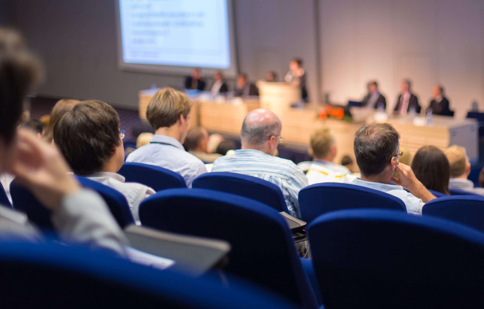 shutterstock_539400208-scaled Individuals sitting in blue chairs at a conference